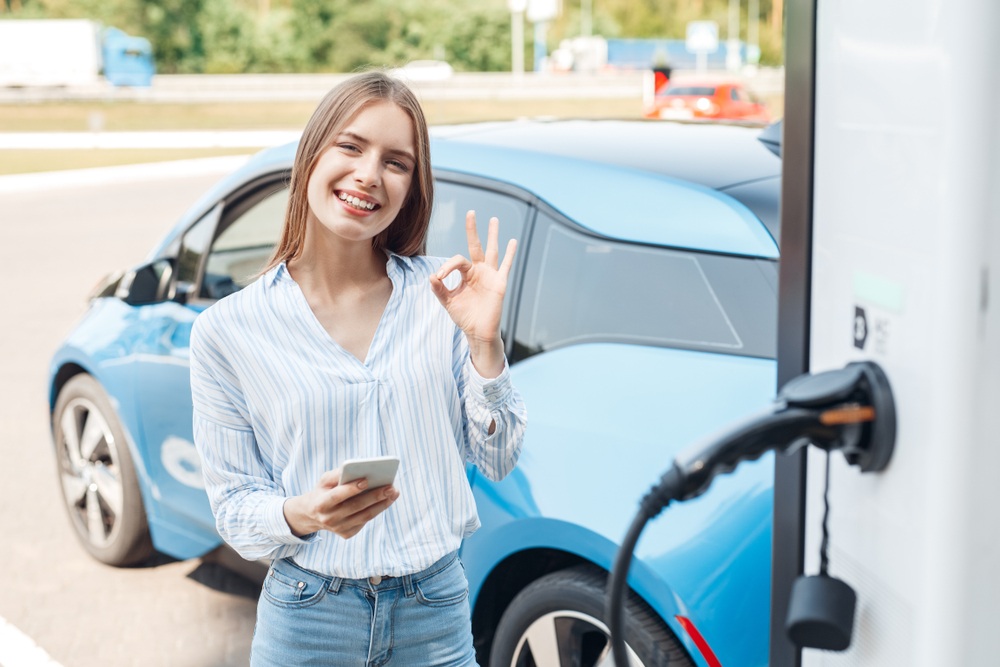 Woman smiling and holding her phone while charging her electric car.