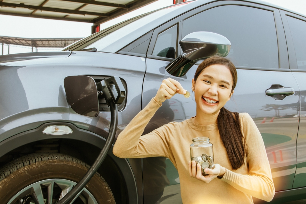 Young Asian woman showing a coin while charging her electric car.